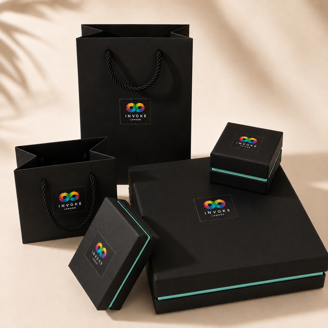 A set of black branded gift packaging from Invoke London, including a large paper shopping bag, a flat gift box, a smaller square box, and a rectangular presentation box, arranged on a warm beige surface with soft leaf shadows.