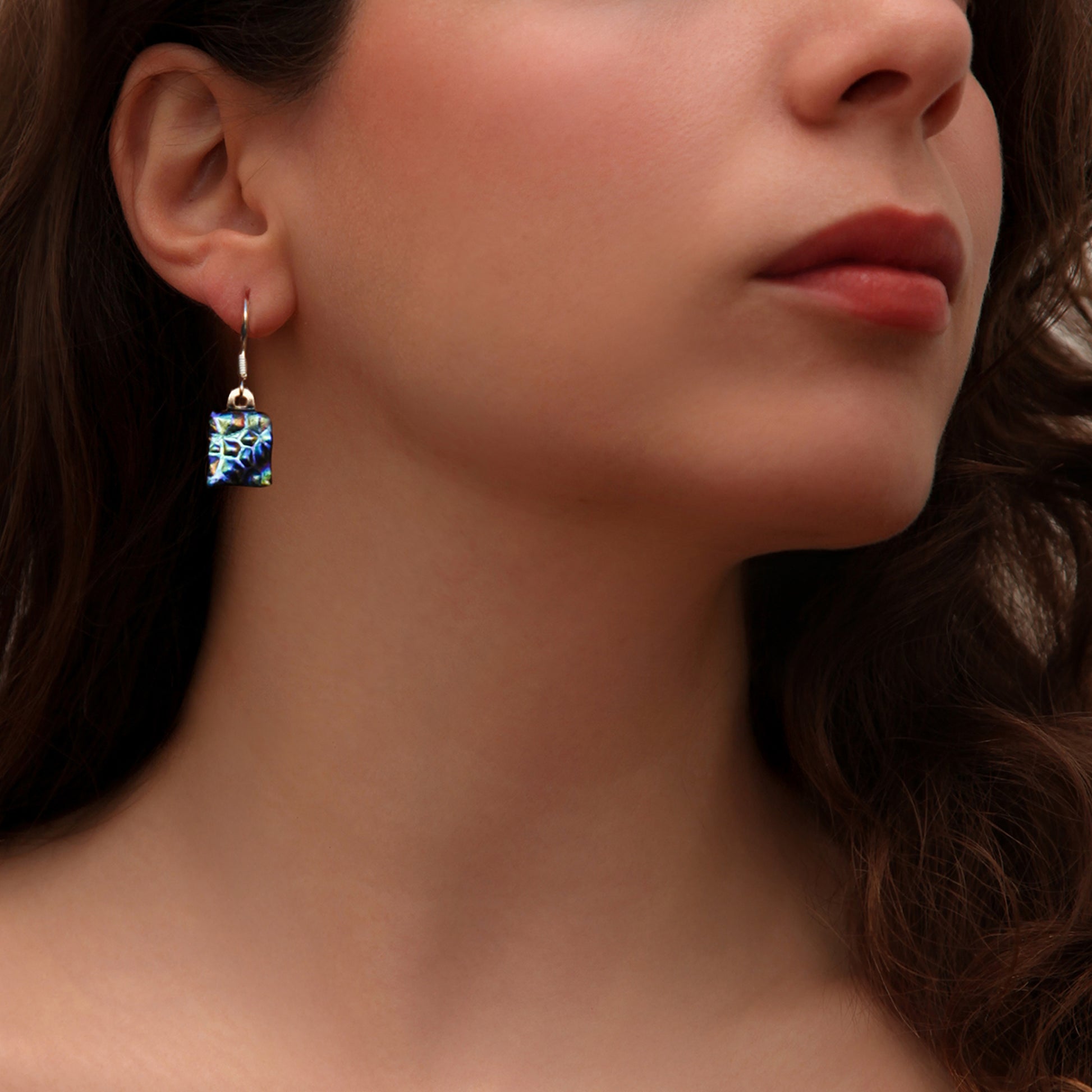 Close-up of handmade dichroic glass drop earring in peacock blue and violet hues – sustainable jewellery by Invoke London.