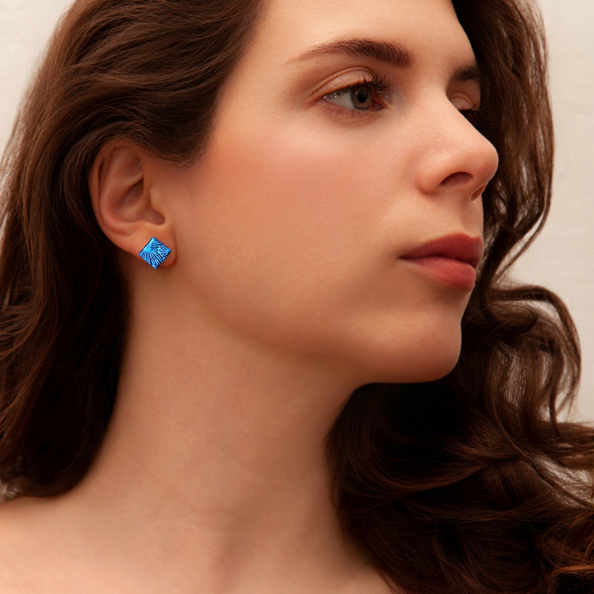 Close-up of woman wearing square blue florentine dichroic glass stud earrings from the Astra collection by Invoke London – handmade in London.