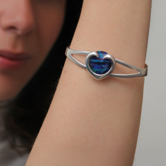 Silver bracelet with a heart-shaped blue, green, purple stone on a wrist.