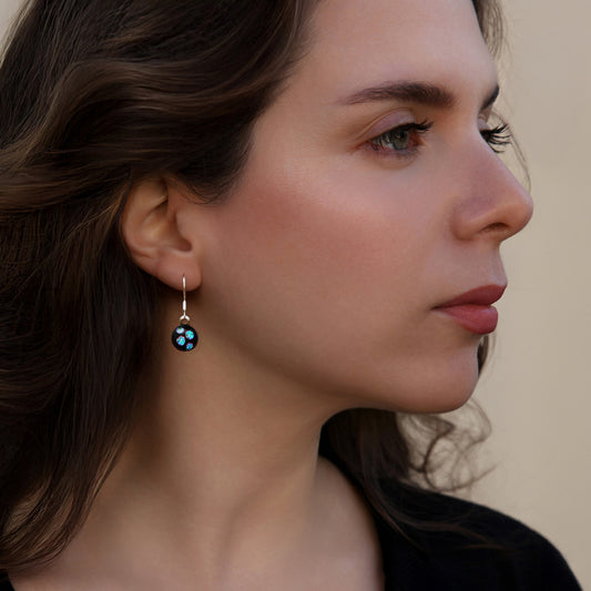 Close-up of model wearing round drop earrings in the Orb Collection by Invoke London, featuring a black fused glass base with shimmering green dichroic dots and silver-tone hooks.
