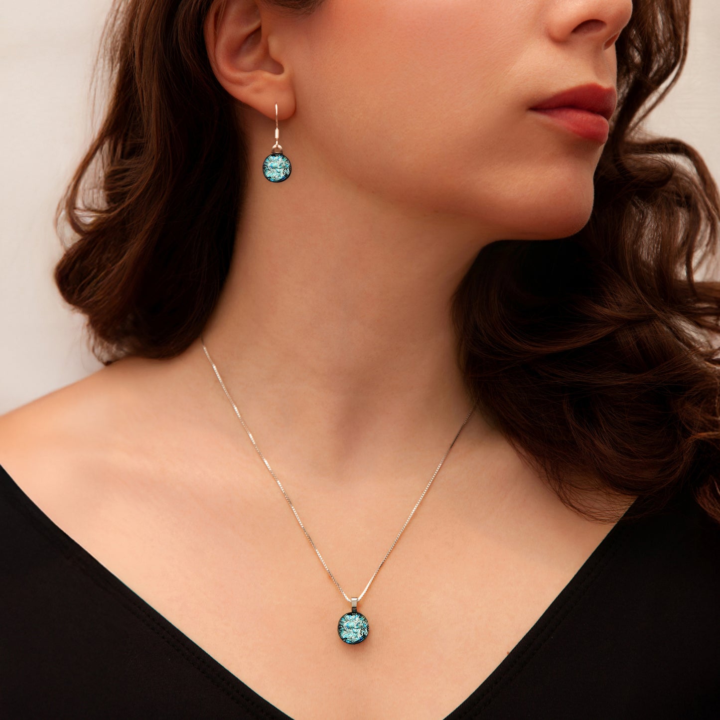 Model wearing a matching jewellery set from the Orb Collection by Invoke London, featuring round silver-toned dichroic glass drop earrings and a coordinating pendant necklace with shimmering aqua and teal tones.