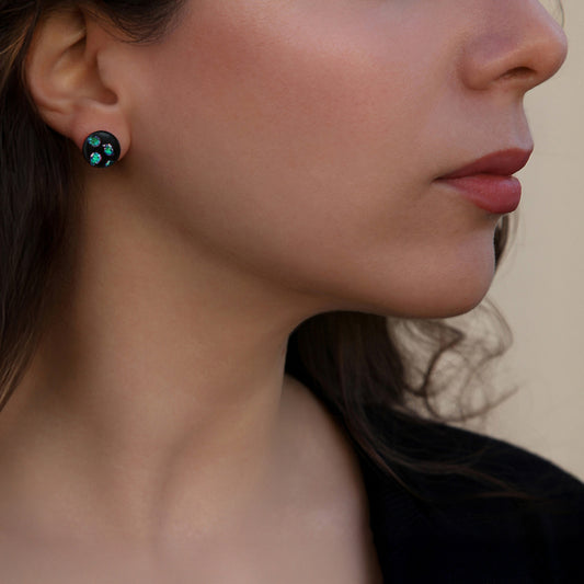 Close-up of model wearing Orb Collection stud earrings by Invoke London, featuring black fused glass with shimmering green dichroic dots.
