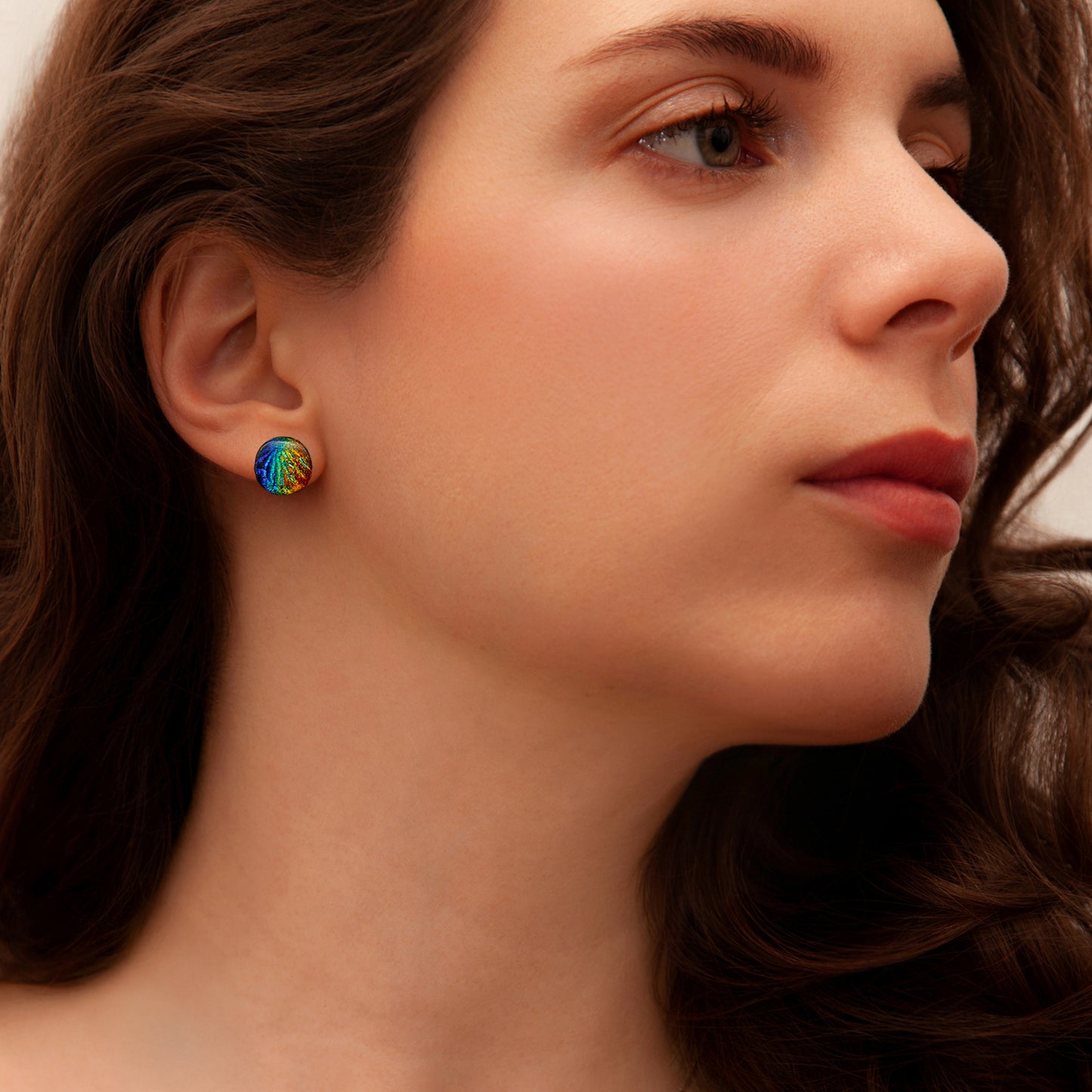 Close-up of model wearing round dichroic glass stud earrings in rainbow tones with a textured Florentine finish, from the Orb Collection by Invoke London.