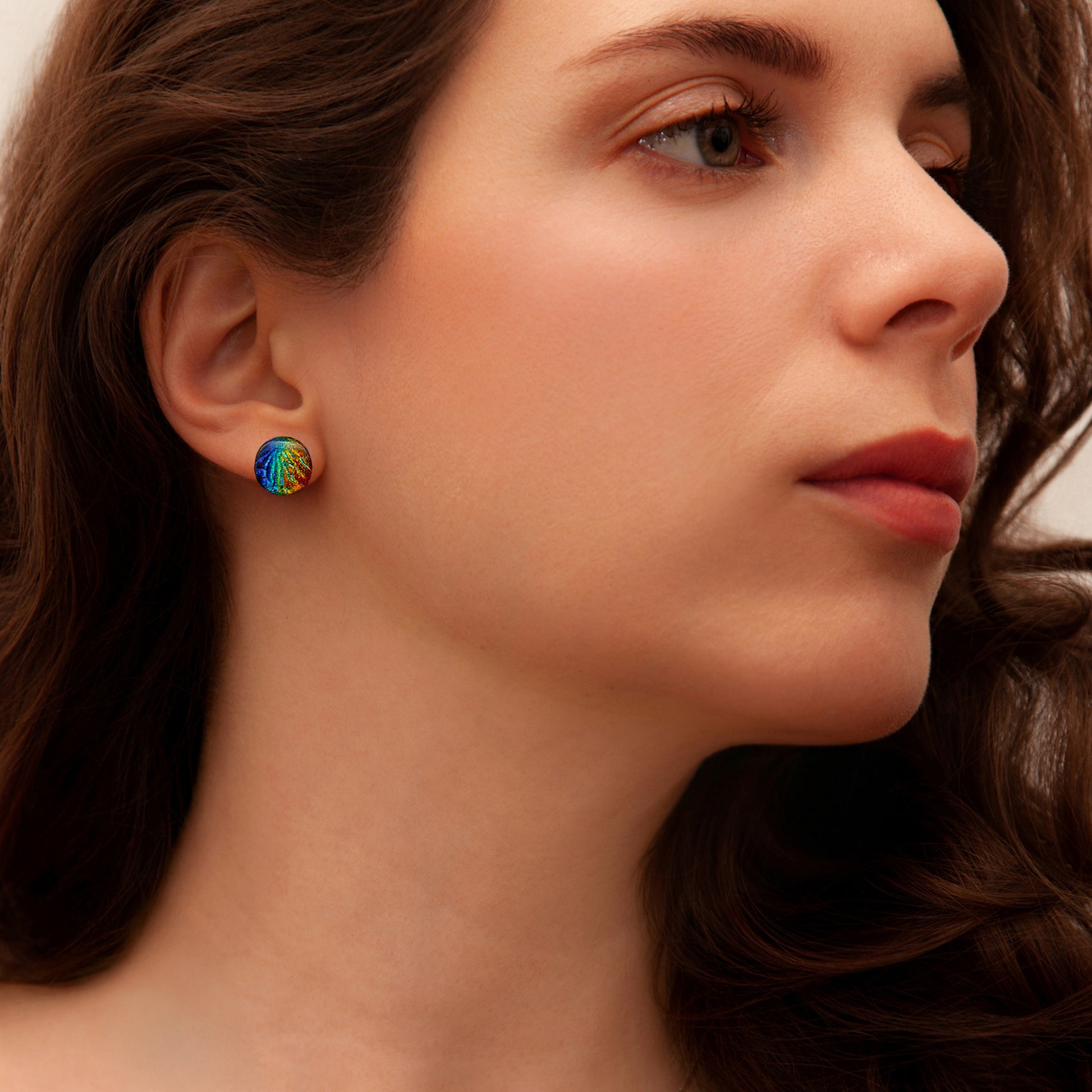 Close-up of model wearing round dichroic glass stud earrings in rainbow tones with a textured Florentine finish, from the Orb Collection by Invoke London.