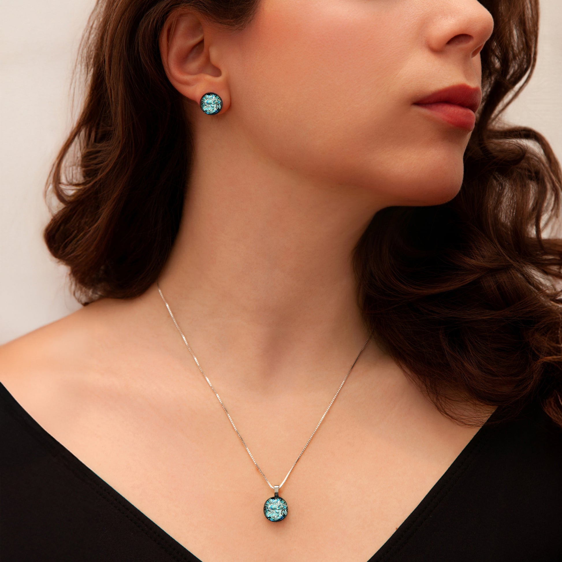 Model wearing a matching jewellery set from the Orb Collection by Invoke London, featuring round silver-toned dichroic glass stud earrings and a coordinating pendant necklace with shimmering aqua and teal tones.