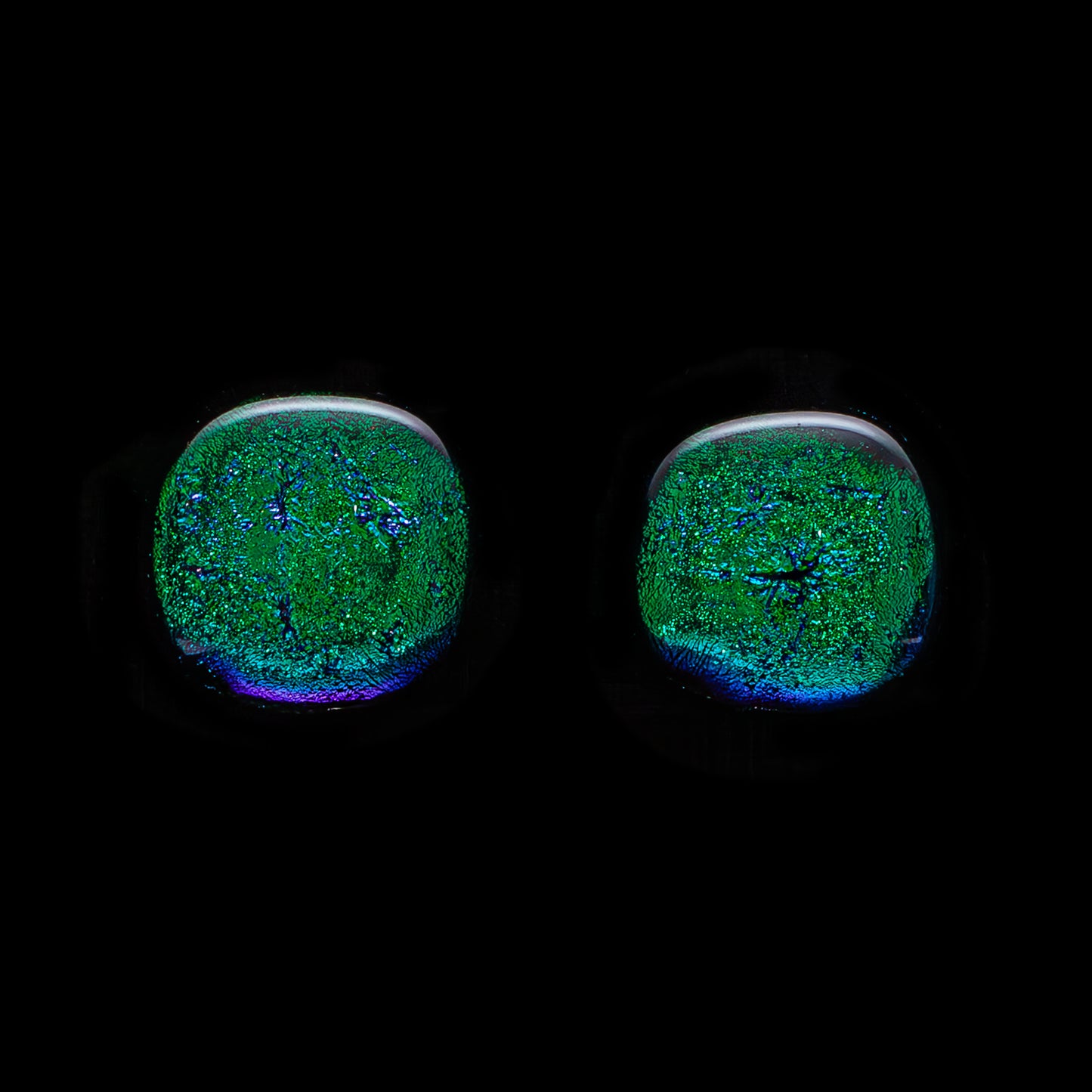 A pair of large, round, dark green dichroic glass stud earrings against a black background.