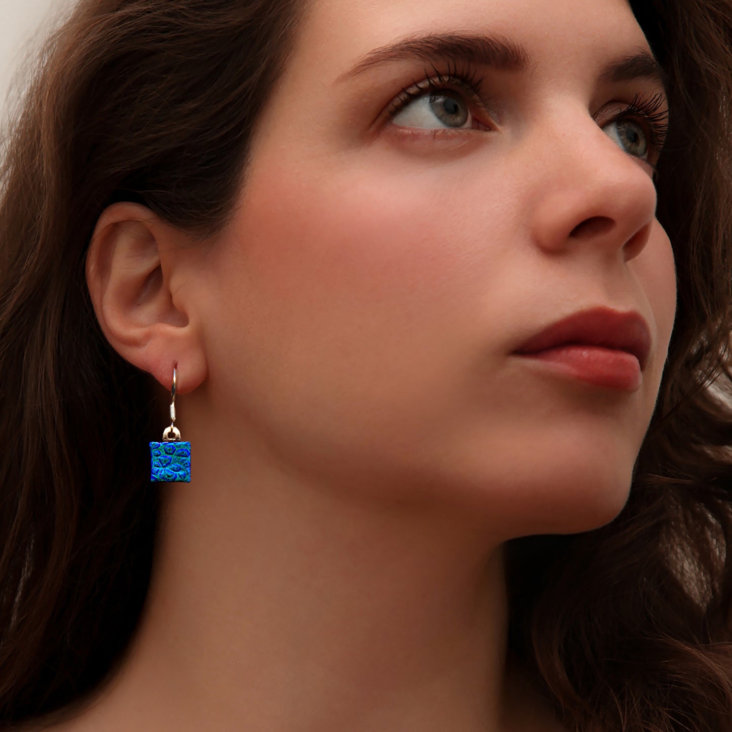 Close-up of woman wearing square blue dew drop dichroic glass drop earrings from the Astra collection by Invoke London – handmade in London.