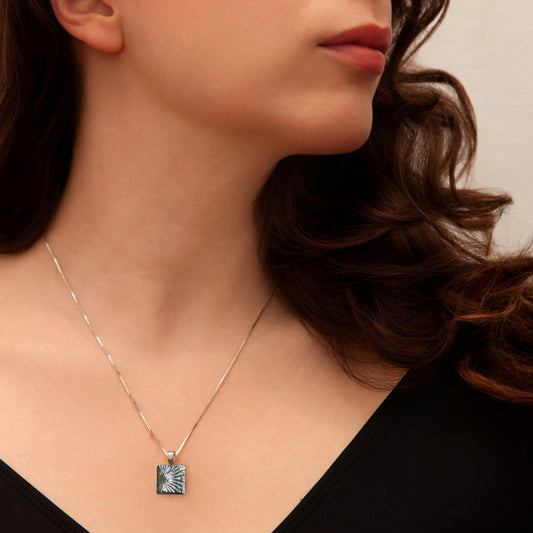 Woman wearing a square silver dichroic glass pendant necklace with sunburst detailing, styled with a black top for Invoke London's Astra collection.