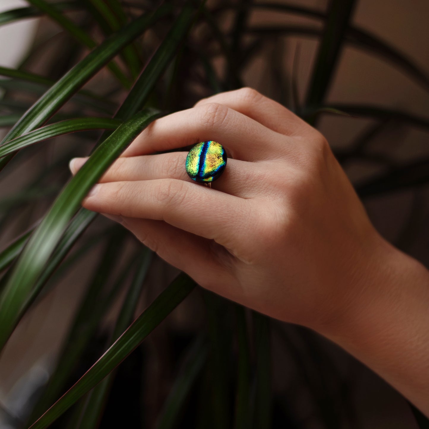 Hand wearing a colorful ring with green and blue stones, held against a plant background