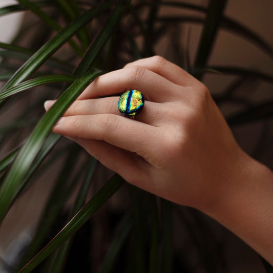 Hand wearing a colorful ring with green and blue stones, held against a plant background